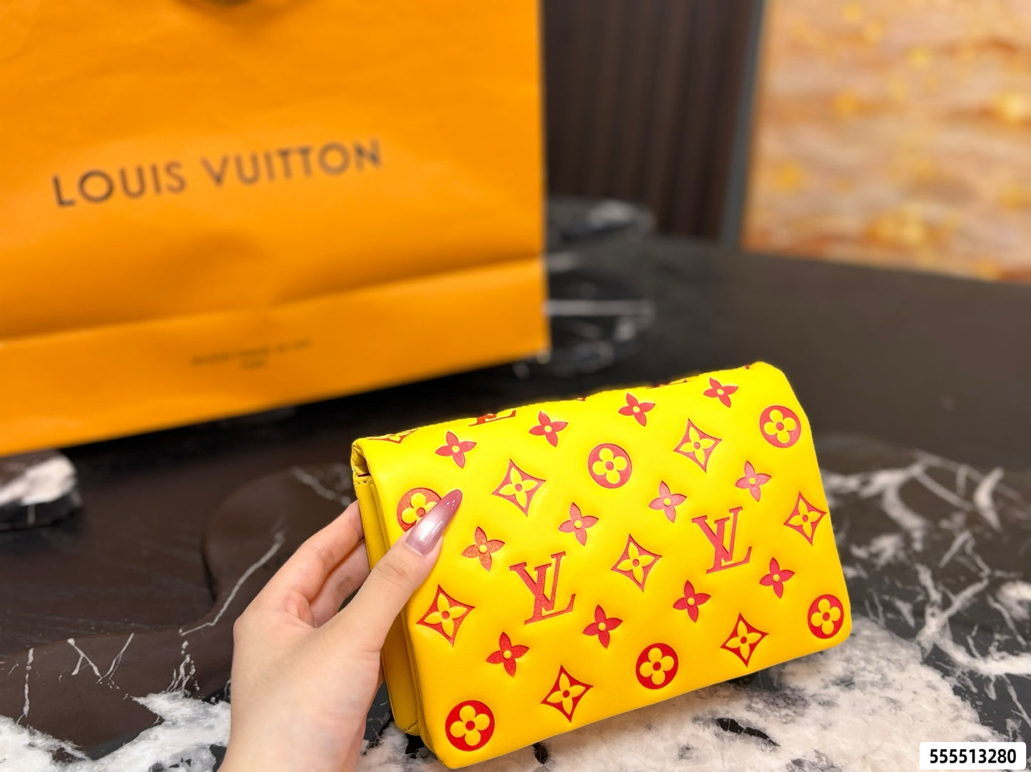 A hand holding a bright yellow Louis Vuitton pouch with the brand's classic monogram pattern, with a blurred orange Louis Vuitton shopping bag in the background.