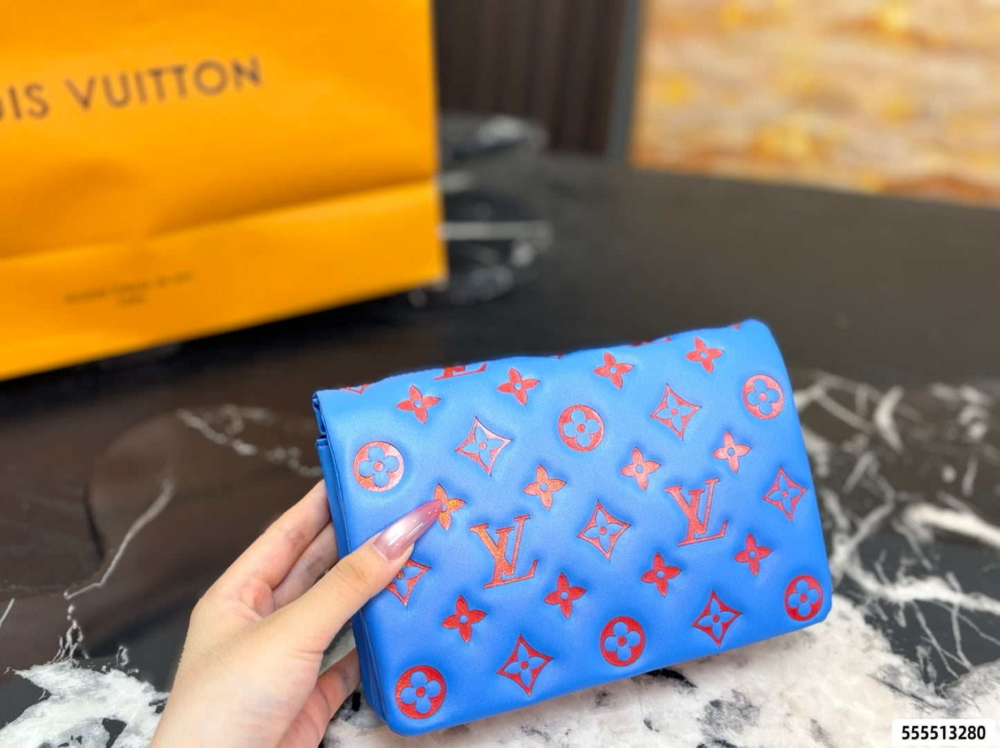 A hand holds a blue Louis Vuitton pouch featuring the brand's signature red monogram pattern. A yellow Louis Vuitton shopping bag is visible in the blurred background on a dark marble surface.