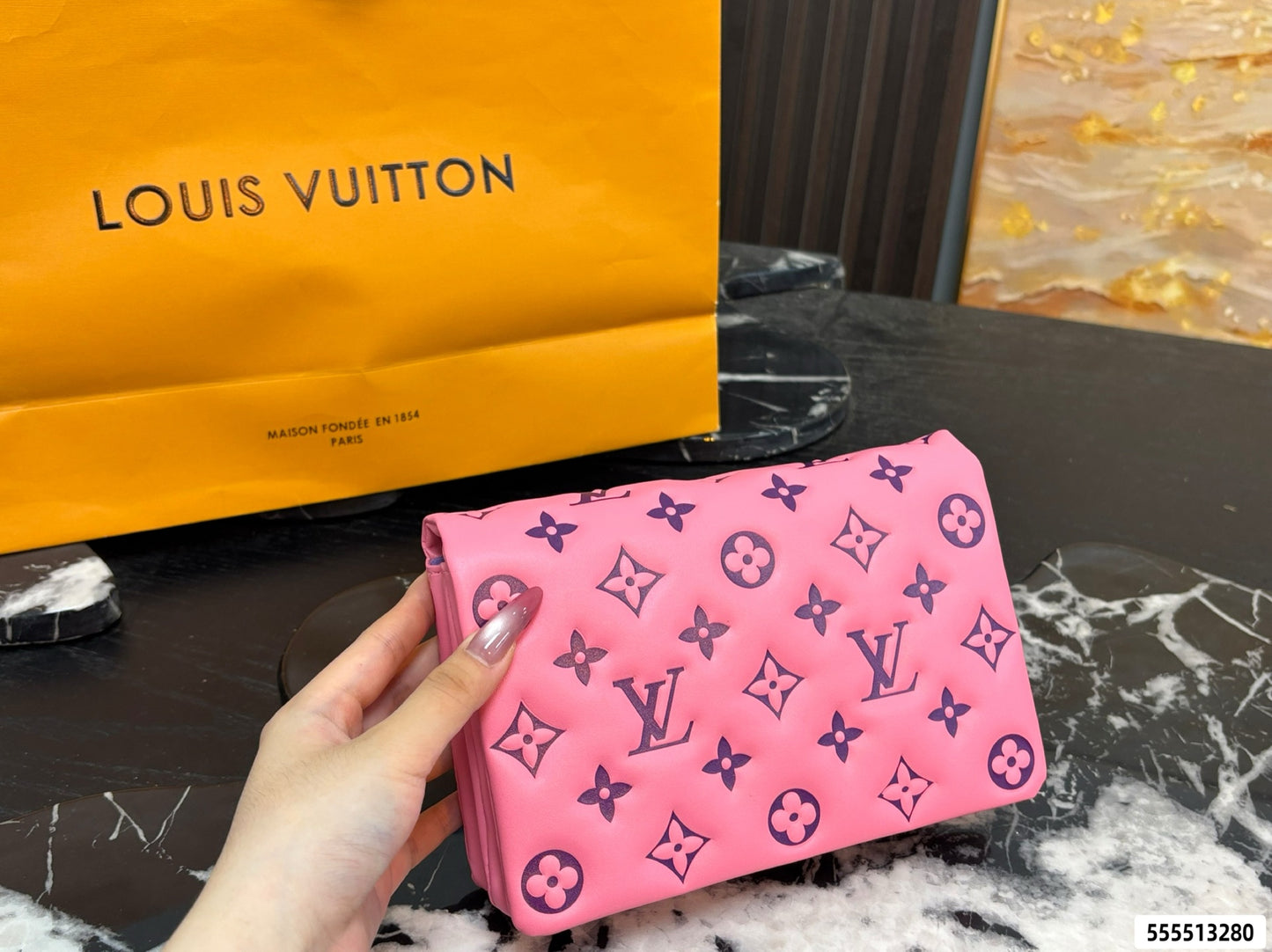 A pink Louis Vuitton monogram wallet or clutch is held in a hand, with a large orange Louis Vuitton shopping bag in the background on a dark marble surface.