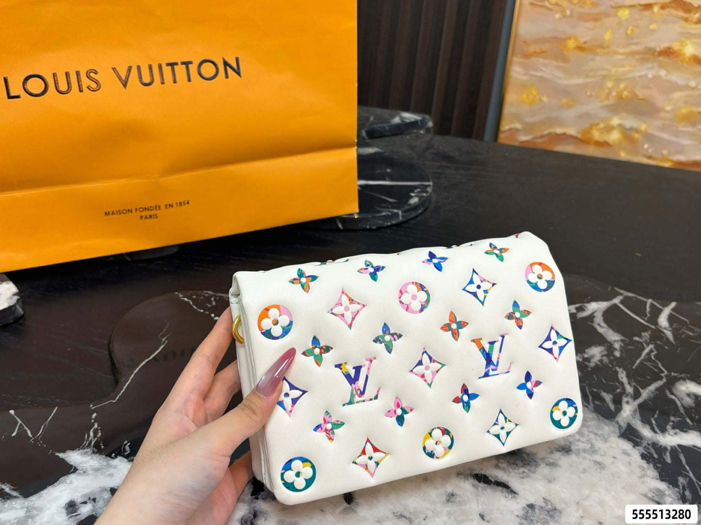 A hand holds a white Louis Vuitton pouch covered in colorful monogram and floral motifs, with a large orange Louis Vuitton shopping bag in the background on a dark marble surface.
