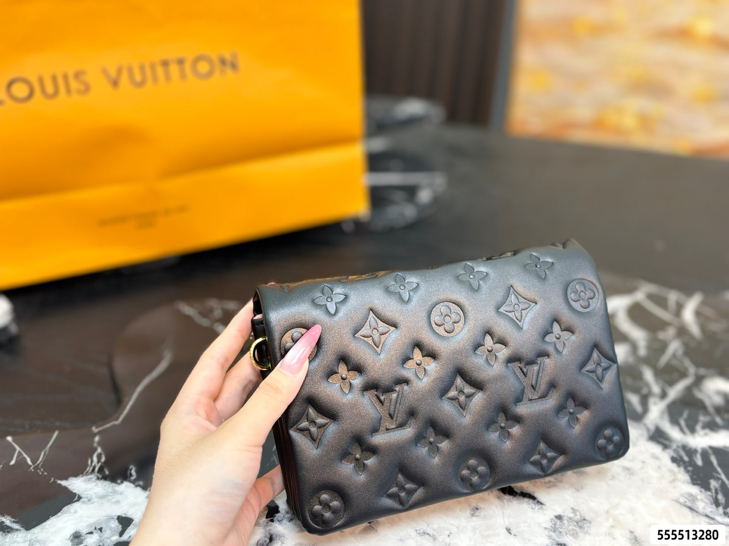 A hand holding a dark brown Louis Vuitton monogram wallet or clutch, with a bright orange Louis Vuitton shopping bag blurred in the background.